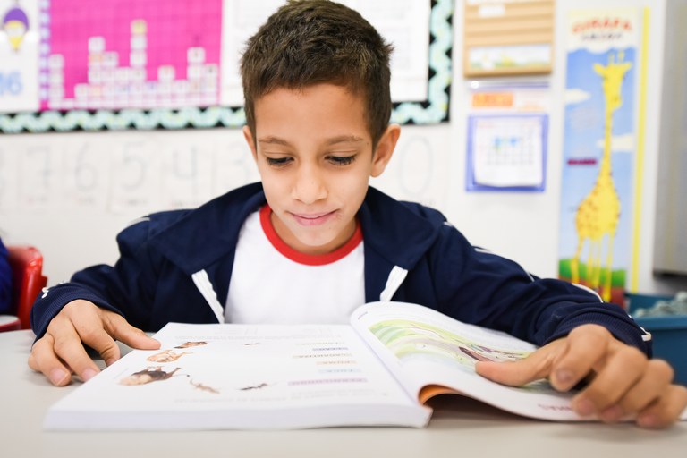 Imagem de um menino com um livro na mesa, estudando na sala de aula